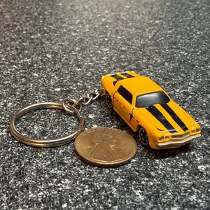 May include: A yellow toy car keychain with two black racing stripes running along the hood and roof. The metal keyring is attached to the rear of the vehicle. A copper-colored United States one-cent coin is placed next to the car for scale.