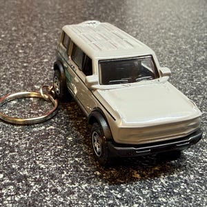 May include: A silver-colored toy SUV keychain featuring a boxy body design, black wheel arches, and black bumpers. The vehicle has a metallic finish, a roof with molded details, and a circular metal split ring attached to the side for keys.