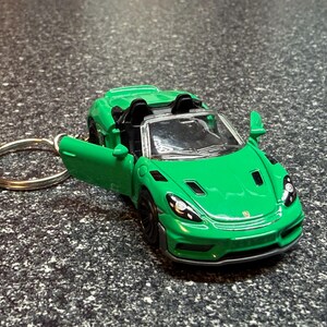 May include: A bright green die-cast sports car keychain featuring an open driver-side door, a clear windshield, black interior seating, and detailed headlights. The miniature vehicle is attached to a standard circular silver metal key ring.