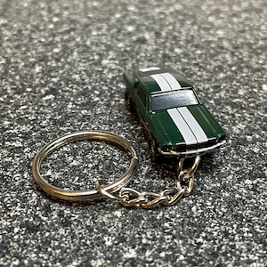 May include: A green toy car keychain with white racing stripes. The miniature car is attached to a silver chain and keyring. The keychain is a miniature replica of a classic car.