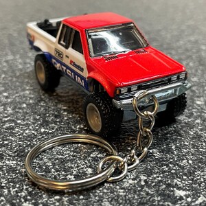 May include: A miniature red, white, and blue Datsun 720 pickup truck keychain. The truck has black wheels and a silver chain and key ring. The truck has the word "Datsun" printed on the side.