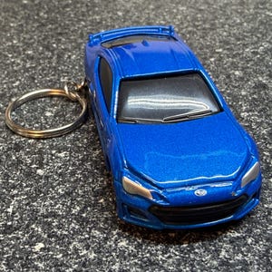 May include: A blue die-cast model car keychain. The miniature car has a metallic finish, with detailed headlights, a front grill, and a Subaru emblem. The keychain ring is silver.