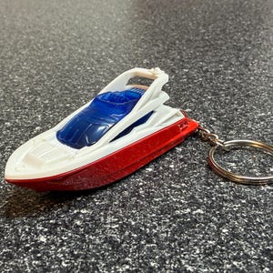 May include: A red, white, and blue toy boat keychain. The boat has a white deck, a blue windshield, and a red hull. The keychain has a silver metal ring.