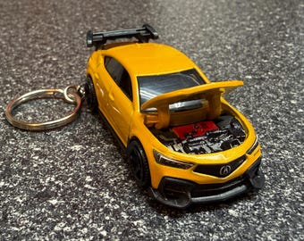 May include: A yellow Acura Integra toy car keychain with a black rear spoiler and black wheels. The hood is open, revealing a detailed engine bay with a red engine cover. The car features the Acura logo on the front grille and clear headlights.