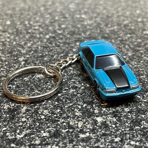 May include: A miniature blue car keychain with a black hood stripe. The car is attached to a silver chain and keyring. The keychain is a small, detailed replica of a classic car.