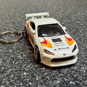 May include: White Toyota 86 diecast model car with a keychain ring. The car features orange and yellow flame graphics, the number 86, and the Goodyear logo. The car has black wheels and a rear spoiler.