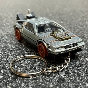 May include: Silver DeLorean keychain with brown wheels and a detailed engine. The miniature car has a chain and key ring attached. The car has the letters "DMC" on the front.