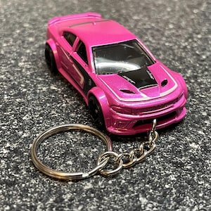 May include: A pink toy car keychain with black accents and a black hood. The keychain has a silver chain and a silver key ring. The car has the word "SRT" on the hood.