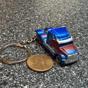 May include: A blue and red semi-truck keychain featuring a metallic finish and silver accents. The truck is positioned next to a copper-colored United States penny, which serves as a scale reference for the size of the miniature vehicle.