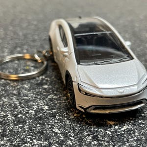 May include: A white toy car keychain with a silver finish. The car has a black interior and a black key ring.