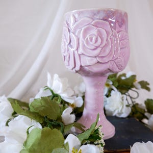 May include: A light lavender ceramic goblet featuring a raised, embossed rose pattern on the bowl and a slender stem. The surface has a glossy, iridescent finish with small star-shaped details integrated into the floral design and stem.