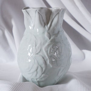 Puede incluir: Jarra de cerámica blanca con un diseño floral en relieve. La jarra tiene un borde festoneado y está decorada con rosas en relieve.