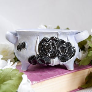 Op de afbeelding: Een lichtlavendelkleurige keramische plantenbak met twee zijhandvatten en drie kleine voetjes. De buitenkant is voorzien van een opvallend, verhoogd reliëf van drie glanzend zwarte bloemen met gladde, ronde bloemblaadjes op een matte lavendelkleurige basis.