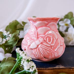 Könnte beinhalten: Eine kleine, glänzende, korallenrosa Keramikvase mit einem erhabenen Reliefdesign, das eine große blühende Rose und eine Biene mit detaillierten Flügeln zeigt. Die Vase hat einen ausgestellten Rand und einen abgerundeten Körper.