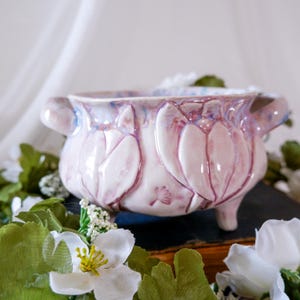 May include: A ceramic bowl featuring a light pink glaze with subtle blue undertones and embossed tulip motifs. The bowl has two side handles, a wide opening, and stands on three small feet, showcasing a glossy finish and a delicate floral design.