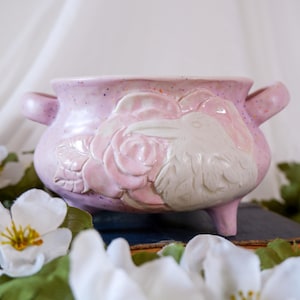 May include: A light pink ceramic cauldron-style bowl with two side handles and three short legs. The exterior features a speckled finish and a raised, embossed relief design depicting a detailed rose flower next to a bird in profile.