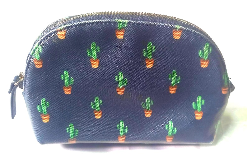 Puede incluir: Un neceser azul marino con un estampado repetido de cactus verdes en macetas naranjas. La bolsa tiene una cremallera dorada y una peque&ntilde;a leng&uuml;eta. La bolsa tiene forma redondeada.