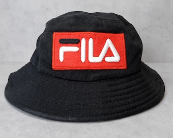 May include: A black bucket hat featuring a rectangular red patch on the front. The patch displays the white embroidered text FILA. The hat has a wide, downward-sloping brim and stitched detailing around the crown and brim for added structure.