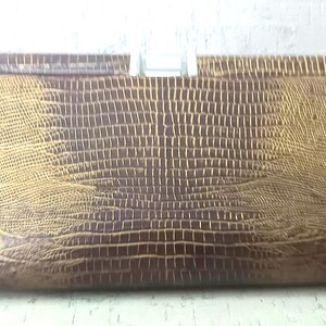 May include: A rectangular clutch bag featuring a brown and gold lizard-skin embossed texture. The bag has a structured, elongated shape with a metallic silver-toned clasp closure centred at the top edge, providing a secure fastening mechanism.