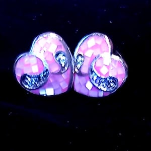 May include: A pair of heart-shaped stud earrings. The earrings are pink with a mosaic pattern and silver detailing. Set against a black background.