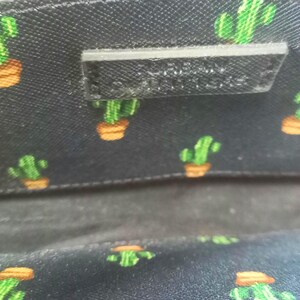 Puede incluir: Primer plano del interior de una bolsa negra con un estampado de cactus. El forro presenta un dise&ntilde;o repetido de cactus verdes en macetas naranjas sobre un fondo oscuro. Se ve una peque&ntilde;a etiqueta negra rectangular.