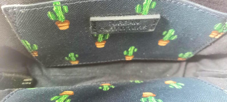 Puede incluir: Primer plano del interior de una bolsa negra con un estampado de cactus. El forro presenta un dise&ntilde;o repetido de cactus verdes en macetas naranjas sobre un fondo oscuro. Se ve una peque&ntilde;a etiqueta negra rectangular.