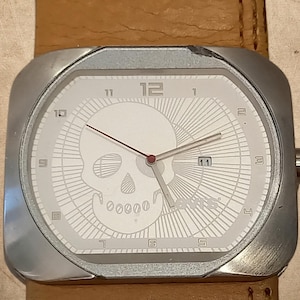 May include: A square-shaped wristwatch with a silver-tone metal case and a tan leather strap. The white dial features a large graphic of a skull, silver-tone hour and minute hands, a thin red second hand, a date window at the three o'clock position, and the Levi's logo.