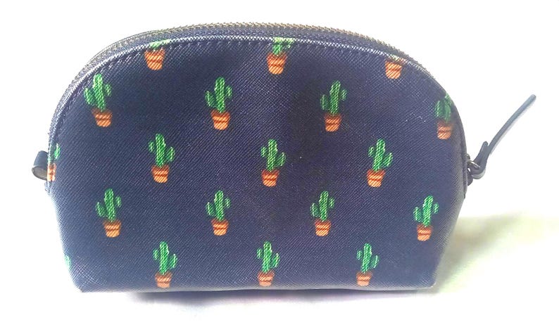 Puede incluir: Un neceser azul marino con un estampado repetido de cactus verdes en macetas naranjas. La bolsa tiene una parte superior curva, un cierre de cremallera y un peque&ntilde;o asa negra.