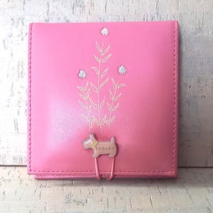 May include: A square pink leather wallet featuring a metallic dog-shaped clasp with the text RADLEY engraved on it. The front surface is decorated with a white line-art illustration of three tall plants and three small circular floral accents.