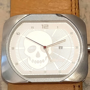 May include: A square-faced wristwatch with a silver-toned metal case and a tan leather strap. The white dial features a prominent skull graphic, silver hour markers, a date window at the three o'clock position, and the Levi's logo in the corner.