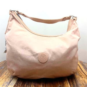 May include: A light pink shoulder bag with a curved design. The bag has a single strap and a small, round emblem on the front. The bag is made of a smooth, possibly nylon material, and has silver-colored hardware.