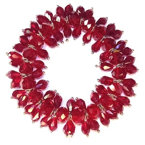 May include: A circular arrangement of faceted, deep red beads. The beads are clustered together, creating a floral-like pattern. The beads are strung on a thin, gold-colored wire. The background is plain white.