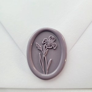 May include: An oval-shaped, light purple wax seal featuring a raised, embossed design of a single iris flower with leaves and a stem. The wax seal is centered on the flap of a plain, white paper envelope, creating a smooth and matte finish.