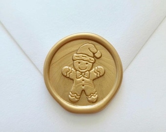 May include: A circular gold wax seal featuring a raised embossed design of a gingerbread person wearing a Santa hat and a bow tie. The wax seal has a smooth, metallic finish and is centered on a plain white envelope flap, creating a festive look.