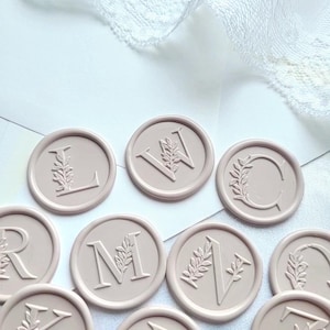 May include: A collection of circular, pale mauve wax seals, each featuring a raised serif letter embossed with a delicate botanical leaf sprig design. The letters L, W, C, R, M, V, and N are visible, arranged on a white surface with lace trim.