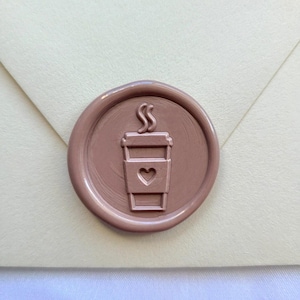 Könnte beinhalten: Ein kreisförmiges, malvenfarbenes Wachssiegel mit einem erhabenen Prägemotiv eines Kaffeebechers, der ein Herzsymbol auf der Vorderseite und aufsteigenden Dampf am Deckel zeigt. Das Siegel ist mittig auf einem cremefarbenen Umschlag platziert.