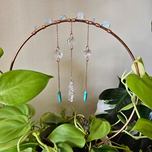 May include: A copper wire hoop with three strands of crystals hanging from it. Each strand has a clear crystal, a clear crystal point, and a turquoise stone. The hoop is hanging from a plant.