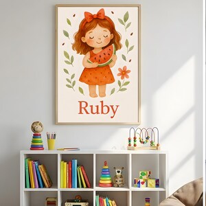 Pode incluir: Uma impress&atilde;o em aquarela emoldurada com uma menina de desenho animado segurando uma fatia de melancia. A menina tem cabelo ruivo e usa um vestido e la&ccedil;o laranja. O nome "Ruby" est&aacute; escrito em vermelho abaixo.