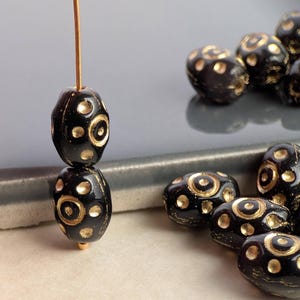 May include: A collection of oval-shaped black beads featuring a gold-colored circular and dotted pattern. Two beads are threaded vertically on a thin wire, while several other identical beads are clustered together in the foreground.