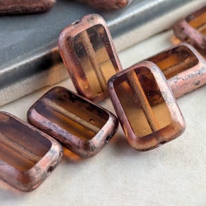 May include: Close-up of several rectangular beads. The beads have a transparent amber center with a copper-colored, textured exterior. The beads are scattered on a white surface, with a gray surface in the background.