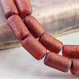 May include: Close-up of a strand of reddish-brown, cylindrical beads. The beads have a textured, mottled appearance with hints of gold. The beads are strung together, suggesting they are for jewelry making or crafting.