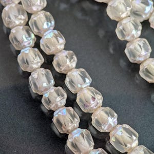 May include: Close-up of strands of iridescent, faceted beads. The beads are clear with a subtle rainbow sheen, and are strung together. The beads have a geometric shape and are arranged in rows against a dark background. The beads are likely for jewelry making.