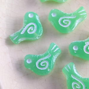May include: A collection of translucent green bird-shaped beads with white spiral and dot details. The beads have a slightly distressed appearance, suggesting an aged or vintage aesthetic. The beads are scattered on a light-colored surface.