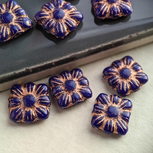 May include: A collection of square-shaped glass beads with a floral design. The beads are a deep blue color with gold accents.
