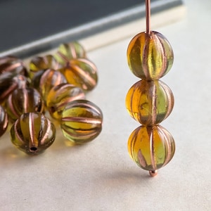 May include: Close-up of several transparent, round beads with a ribbed design and copper-colored stripes. The beads are a mix of yellow and green hues, strung together on a copper wire. Some beads are scattered nearby.