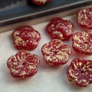 May include: Close-up of several red flower-shaped beads with gold accents. The beads have a textured surface and a hole in the center for stringing. The beads are likely for jewelry making or other craft projects.