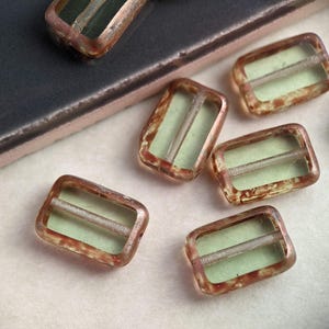 May include: A collection of six rectangular glass beads with a brown and green mottled pattern. The beads have a clear center with a thin, opaque green line running through the center.