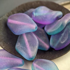 May include: A close-up of a group of iridescent glass beads in shades of blue and purple. The beads are smooth and have a frosted finish. They are arranged in a pile on a dark surface.