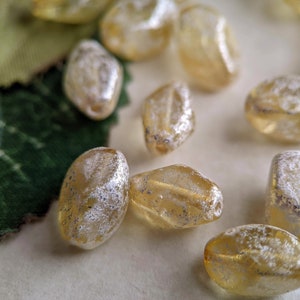 May include: A collection of oval-shaped glass beads with a frosted, iridescent finish. The beads are a light amber color with a subtle shimmer.