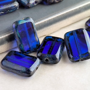 May include: A collection of rectangular, dark blue glass beads with a glossy finish. Each bead has a hole for stringing and features internal striations of lighter blue and green. The beads are arranged on a light surface.